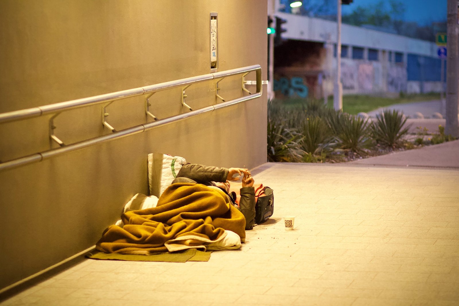 A person is lying on the ground under a yellow blanket in an outdoor walkway, with a cup placed nearby. The scene takes place near a building with a rail and a sidewalk in the background.