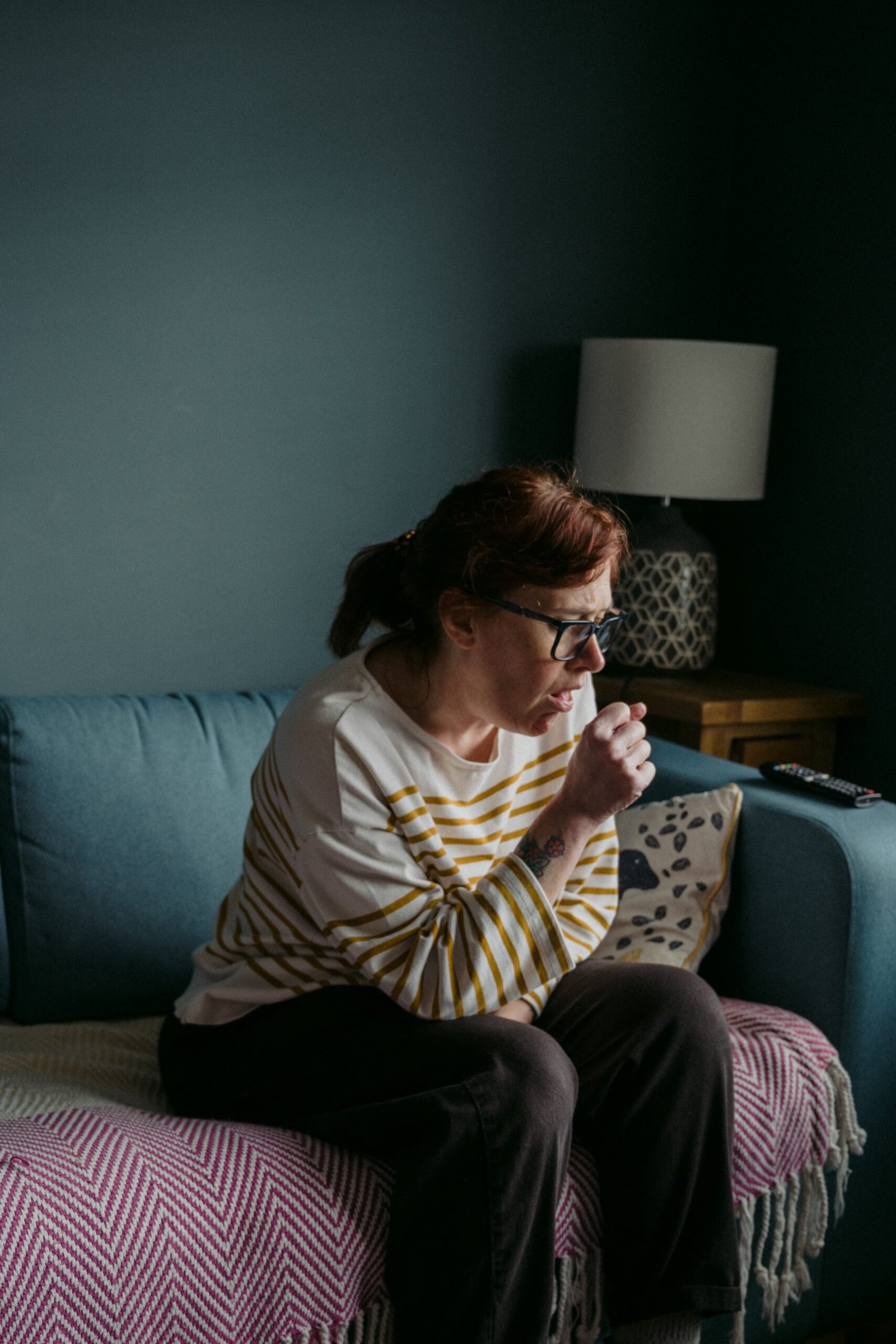 A woman wearing glasses and a striped sweater sits on a sofa, coughing into her fist in a room with a blue wall, a lamp, and a remote control visible.