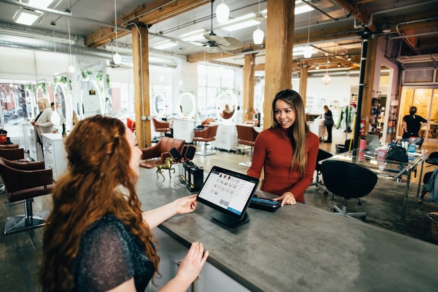 A woman stands at a counter in a salon, talking to another woman who is seated. A tablet with a sales interface is on the counter. Various salon chairs and mirrors are visible in the background.