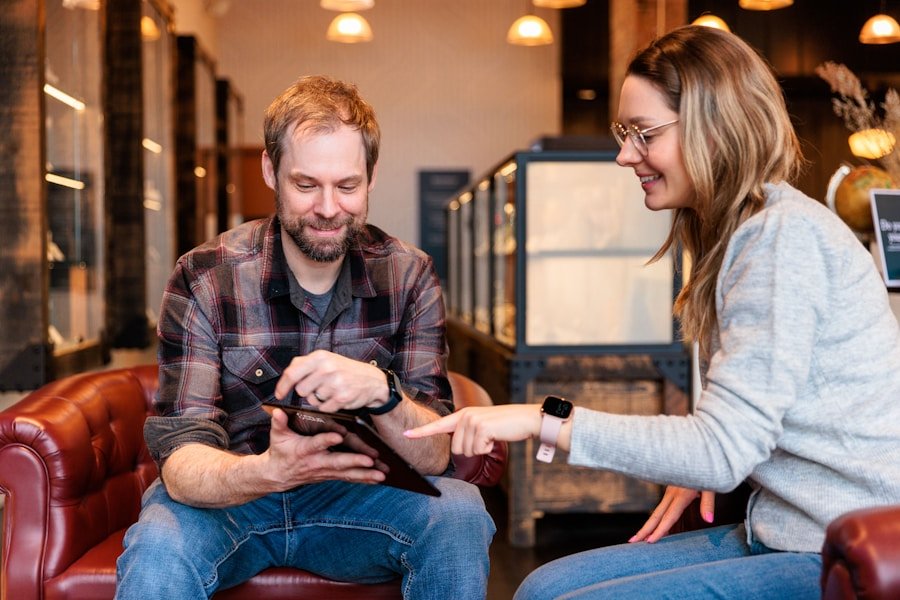 Two people sitting on red sofas, smiling and interacting with a tablet. The man is holding the tablet while the woman points at the screen. Background shows a softly lit cafe or lounge setting.
