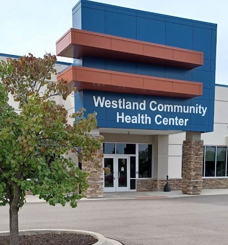 The entrance of Westland Community Health Center, with its glass doors and blue and orange facade accents, reflects our mission of providing specialized care. A tree stands proudly in the foreground, symbolizing growth and healing.