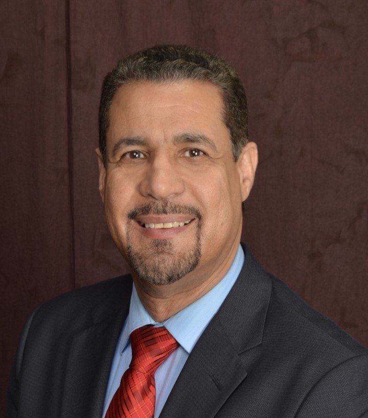 A man in a suit with a red tie and light blue shirt, smiling in front of a brown background, embodies the values of specialized care.